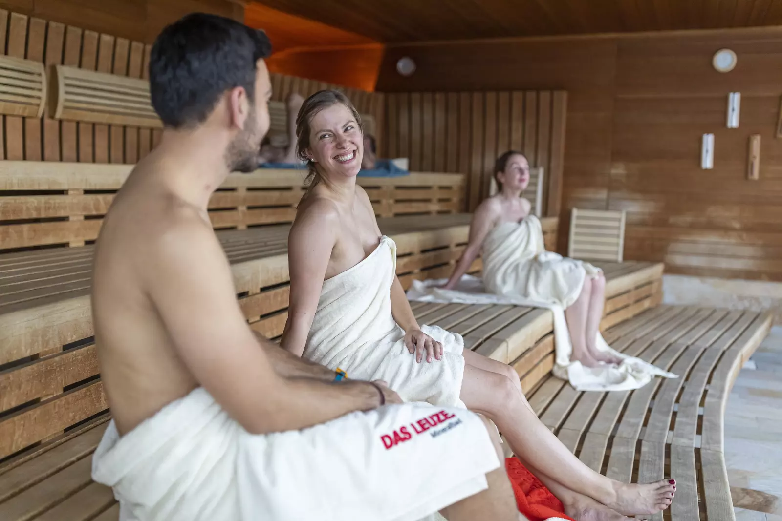Enspannung in der Panorama-Sauna Ein Paar, in Badetücher gehüllt sitzt auf einer Holzbank in der Panorama-Sauna und unterhält sich entspannt und fröhlich.