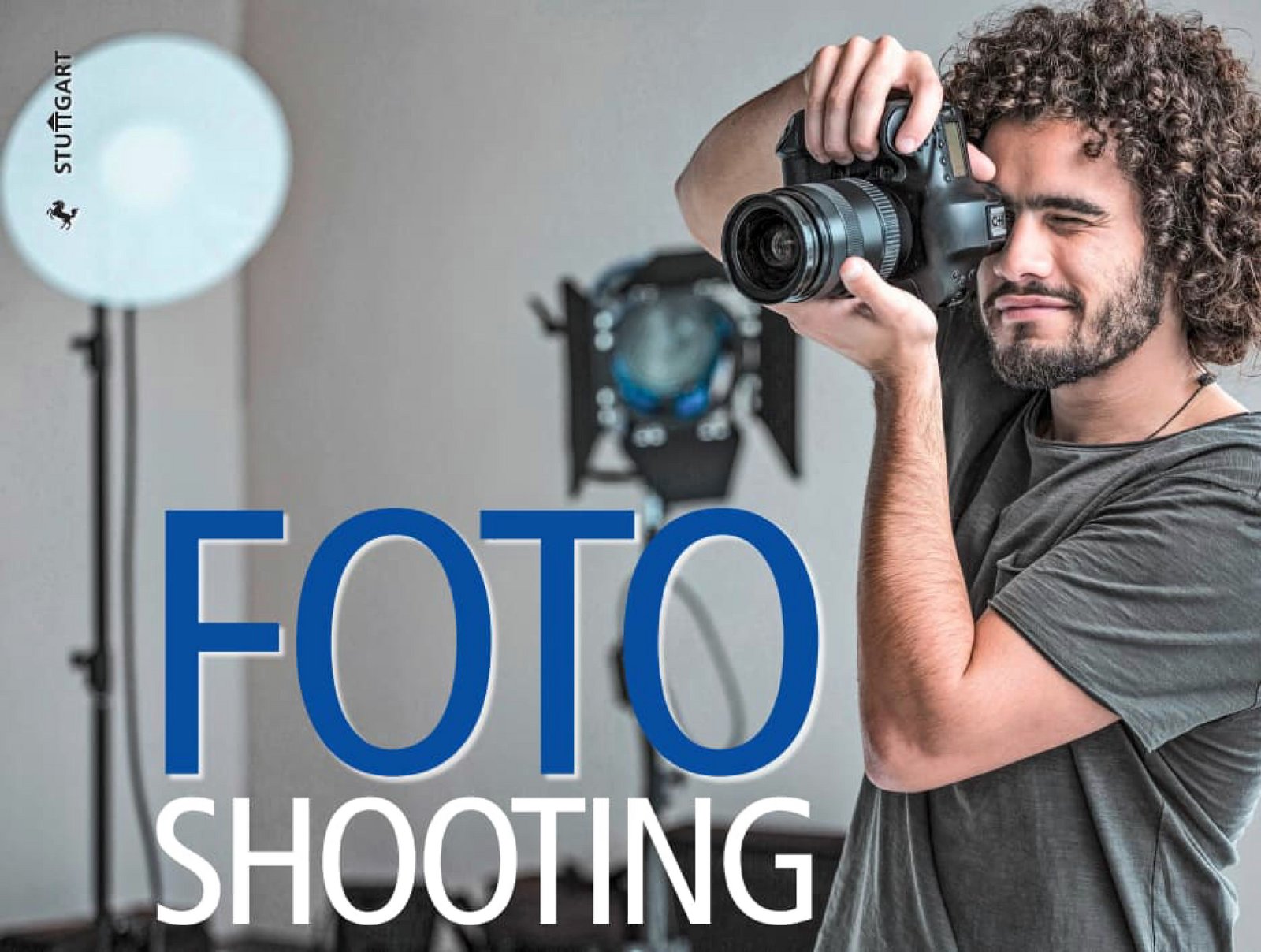 Ein Fotograf hält eine Kamera in den Händen zum fotografieren bereit. Im Hintergrund ist die Fotoausrüstung wie Blitzschirm und Lampe mit Staiv zu sehen.