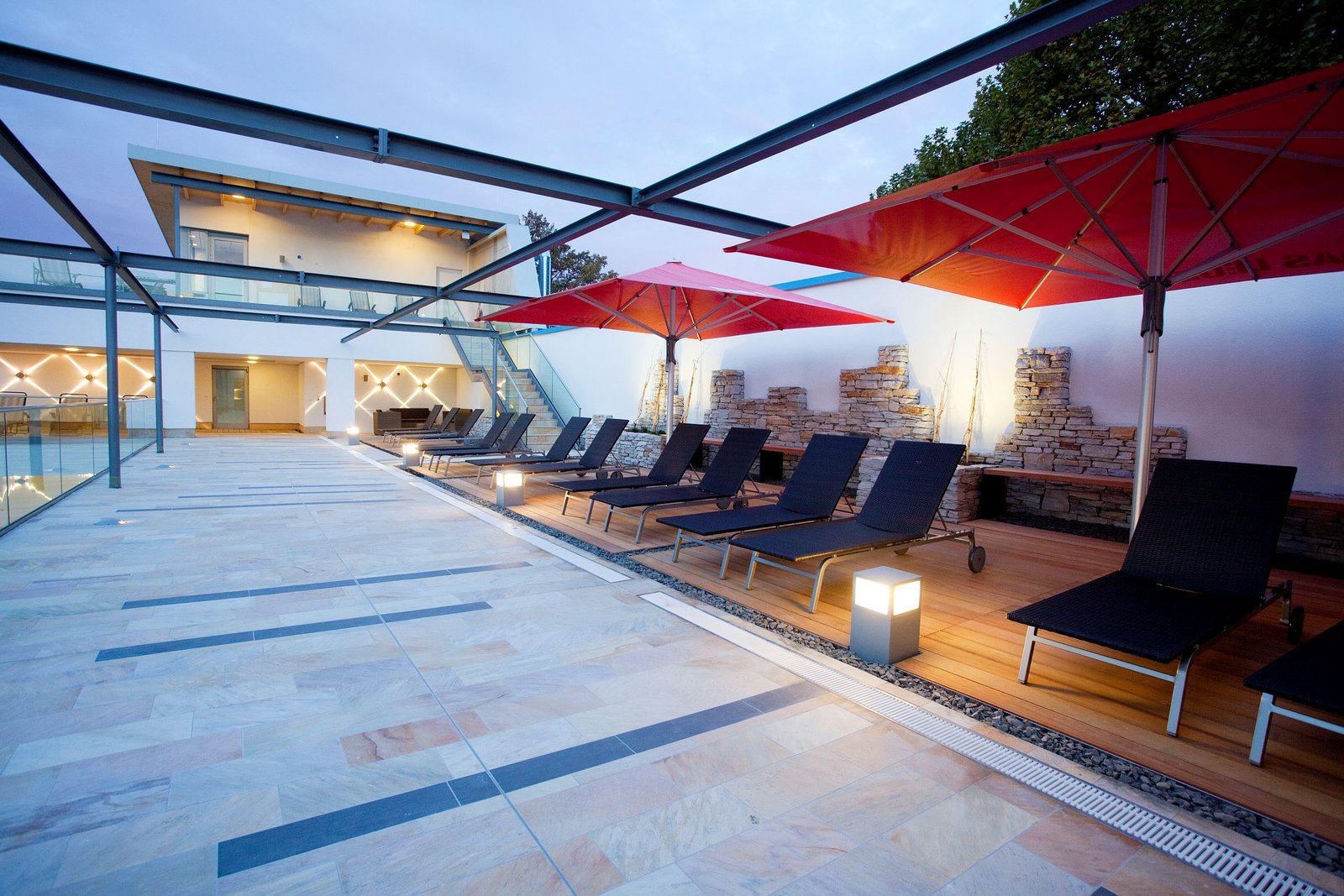 Sonnenterasse mt grauen Liegen und roten Sonnenschirmen. Das Bild ist bei Dämmerung aufgenommen. Sanfte Beleuchtung durch die kleinen Lampensäulen.